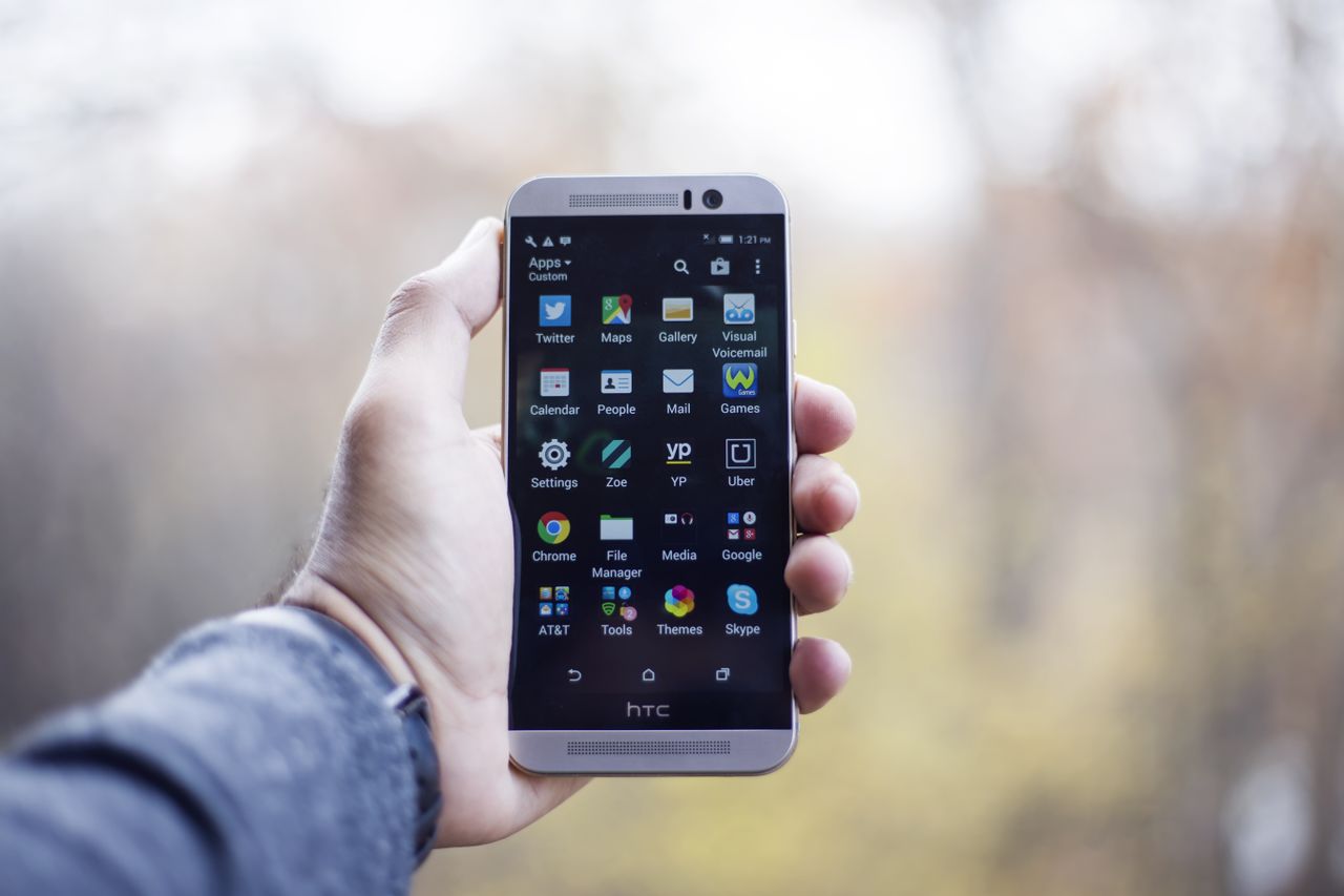 about-img A close-up of a hand holding an HTC smartphone with visible apps on the screen, in an outdoor setting.